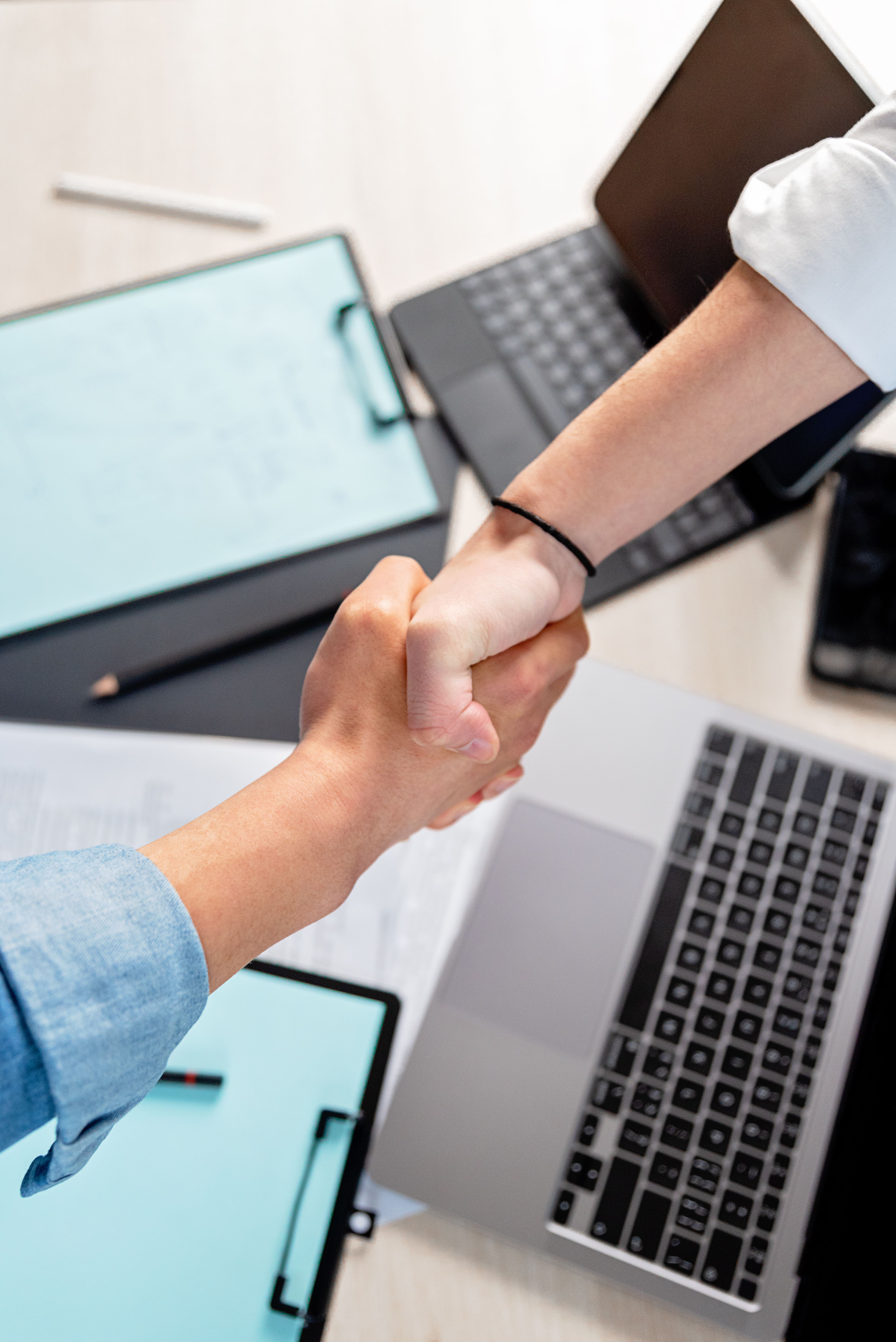Twee mensen geven elkaar een hand boven een bureau met laptops en klemborden.
